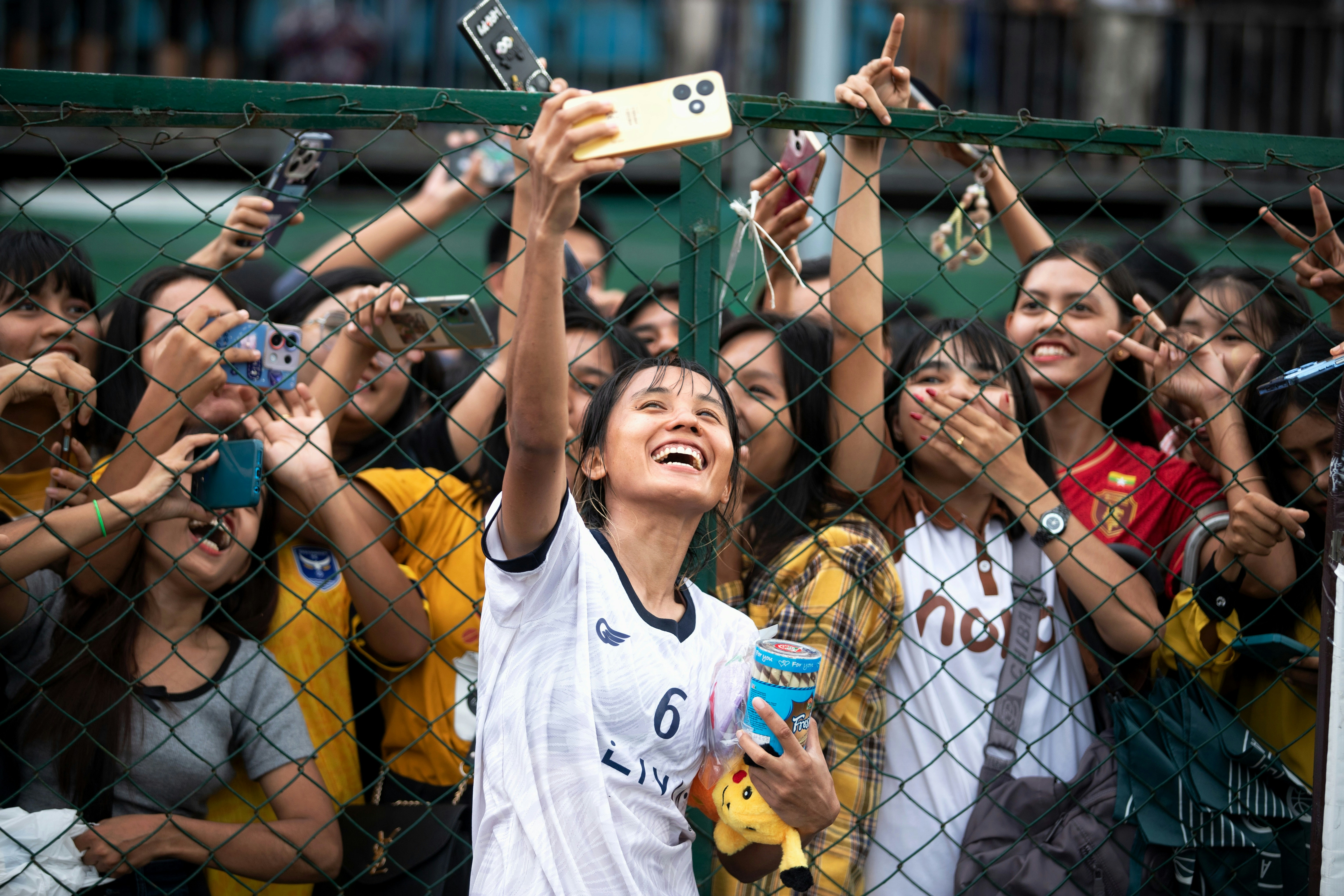 A vibrant moment of pure joy and connection. A young female footballer in Myanmar smiles broadly while taking a celebratory selfie with a crowd of enthusiastic fans behind a fence after a match. Their shared laughter, smartphones in hand, embodies the spirit of optimism, sportsmanship, and community .
